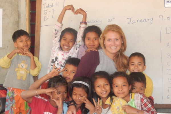 Volunteer teacher and students (1) A woman stands with children, smiling, in front of a whiteboard filled