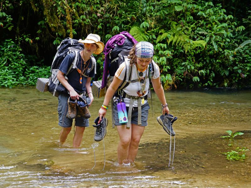 Two individuals with backpacks navigate across a stream