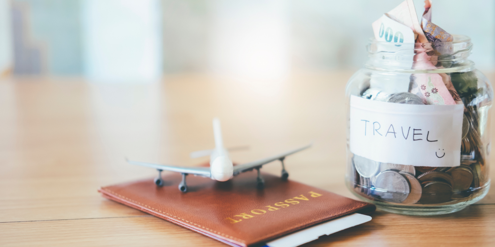 A clear glass jar labeled "TRAVEL" with coins and banknotes inside, next to a miniature airplane on top of a brown passport holder, all sitting on a wooden surface suggesting savings for future travels.