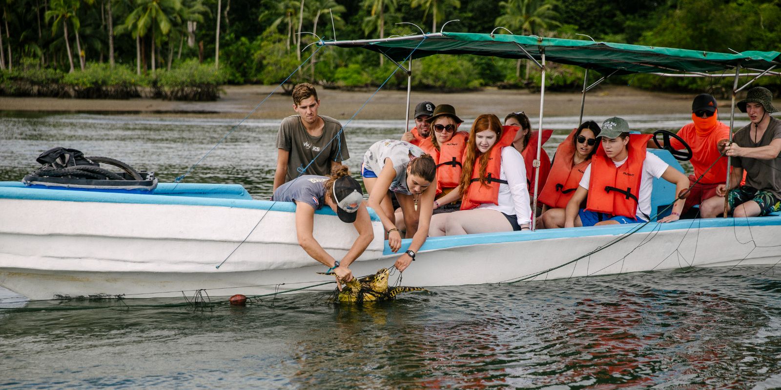 A group of young adults on a boat wearing life jackets, actively engaged in a river cleanup, with tropical vegetation in the background.