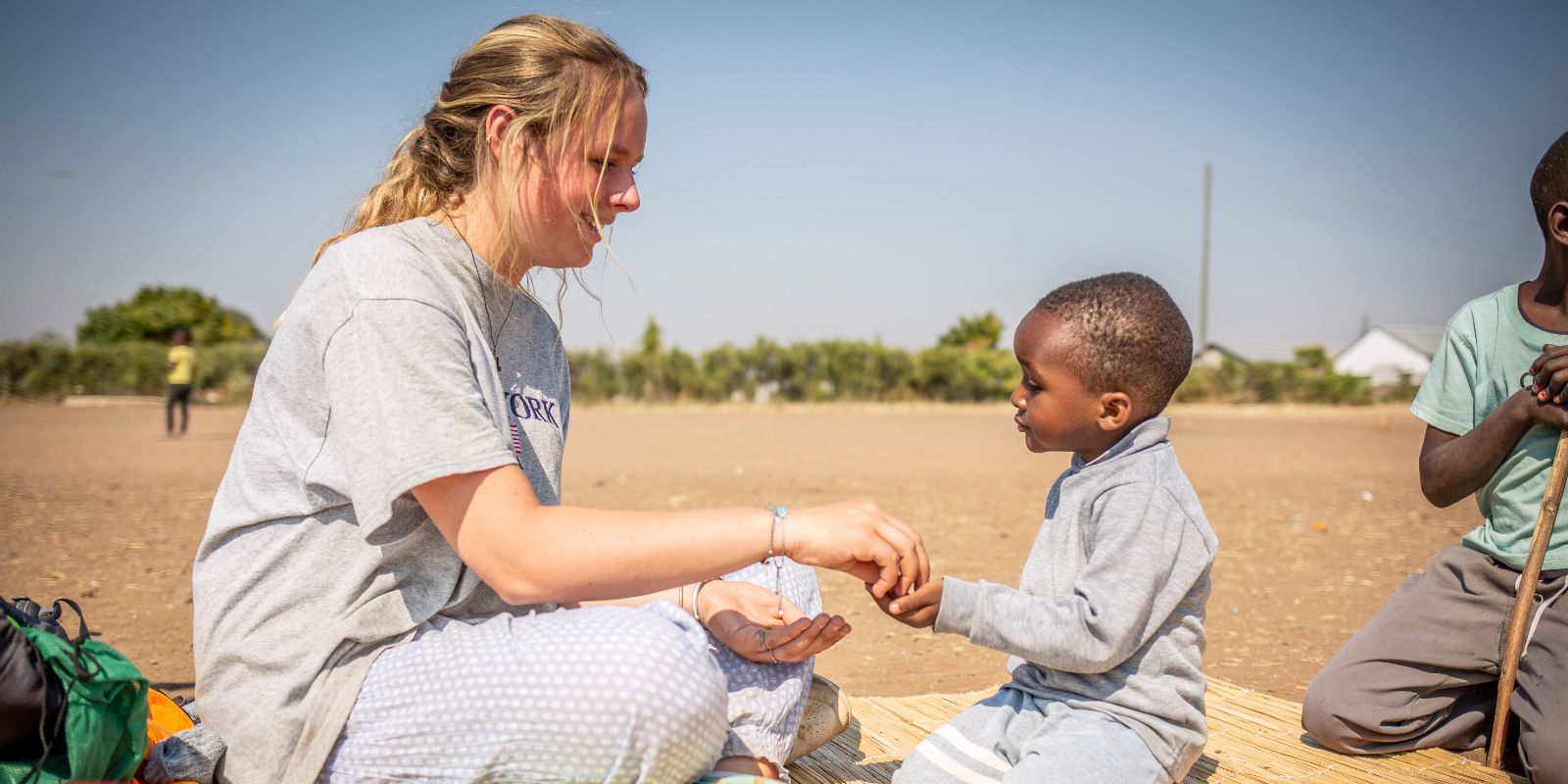 A woman in a grey shirt plays hand games with a young boy on a woven mat outdoors, both smiling in a sunny, rural setting.