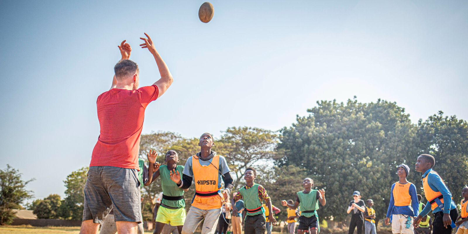 A group of people, likely a mix of locals and visitors, engaged in a game of rugby in an outdoor setting, with a clear focus on interaction, teamwork, and community participation.