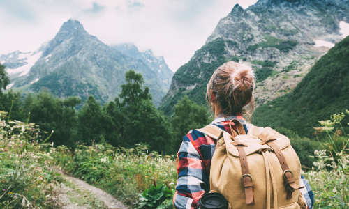 A woman standing before a mountainous landscape, looking into the distance with her back to the camera, wearing a plaid shirt and a backpack, possibly indicating a journey or adventure ahead.