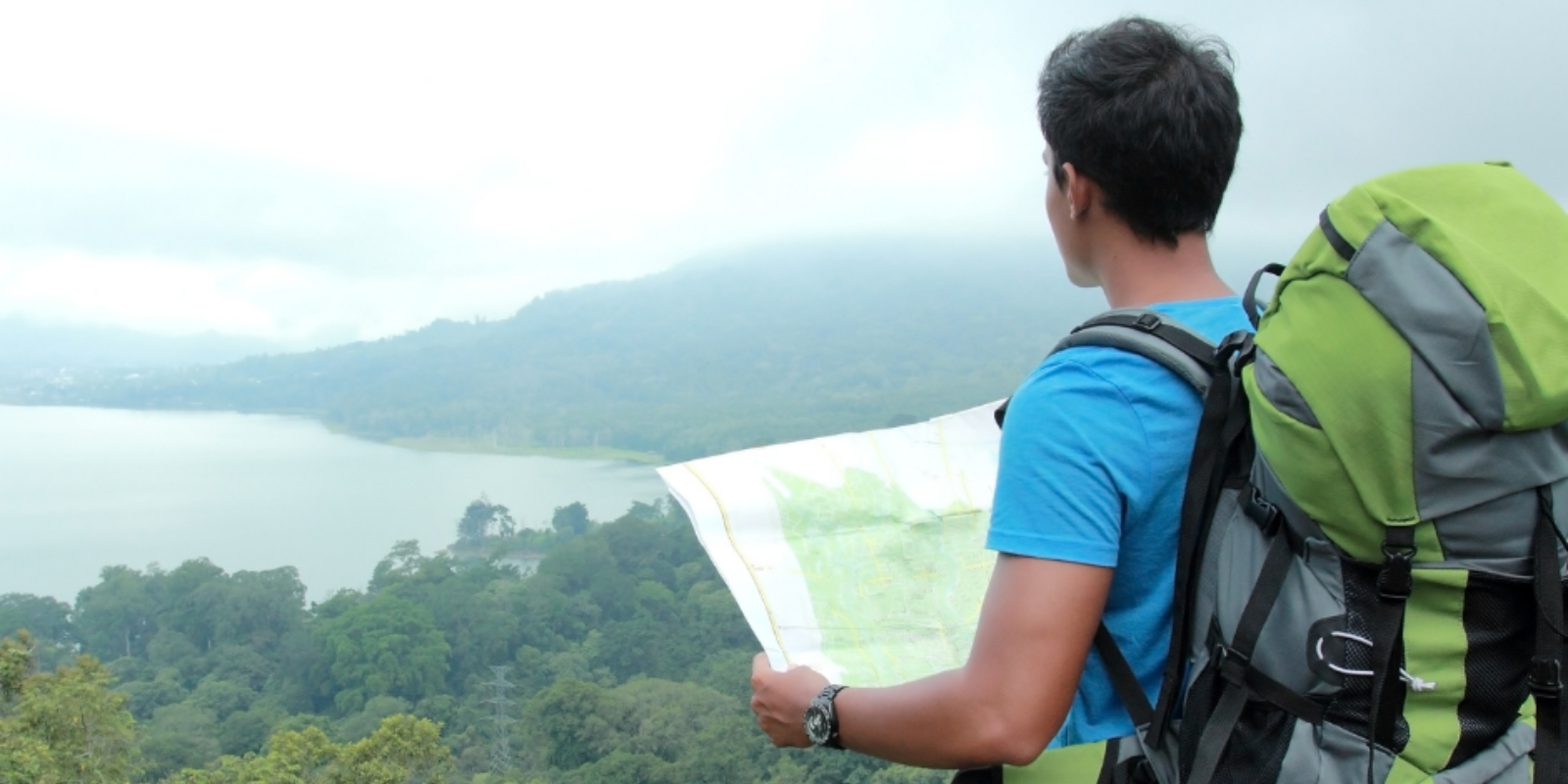 A traveler with a backpack is looking at a map with a scenic lake and lush hills in the background.