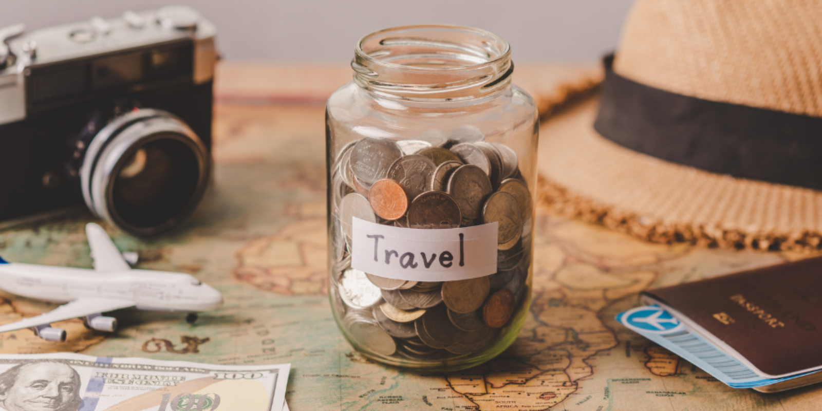 A jar full of coins labeled "Travel" on a map, suggesting saving money for travel, with a vintage camera, a straw hat, a model airplane, and a passport, alluding to planning and preparing for a journey or vacation.