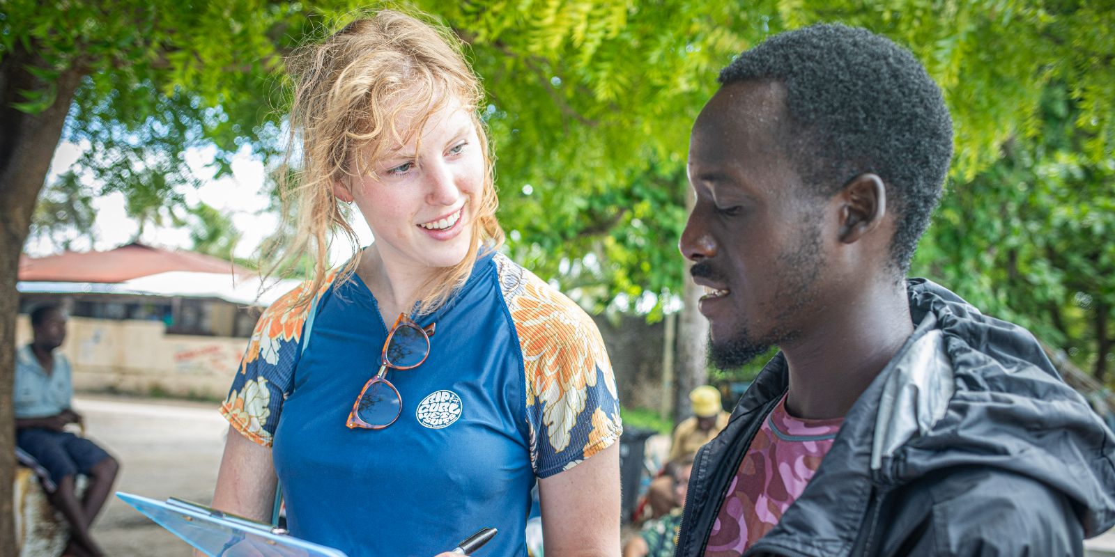 A young woman with blonde hair, smiling as she looks at a clipboard, is in conversation with an African man. They are outdoors, with greenery and a seated person in the background.