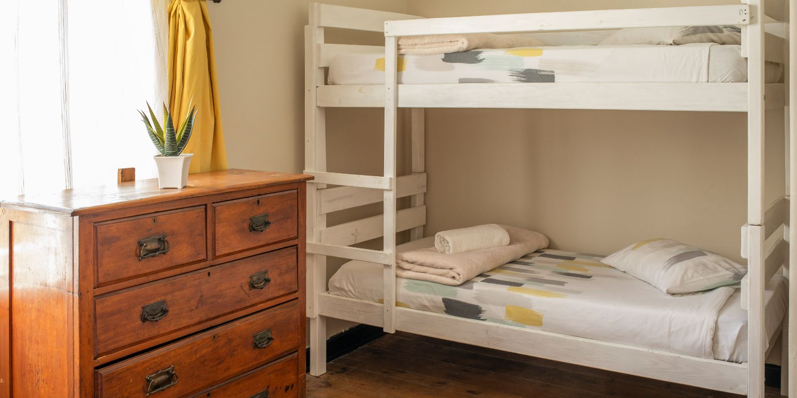 A cozy dormitory room featuring a wooden double-decker bunk bed, a vintage dresser, and a small potted plant on a wooden table near a window with yellow curtains.