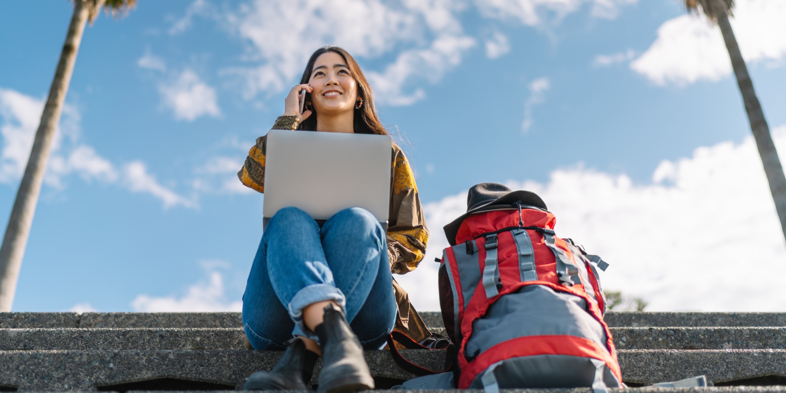 A young woman sits on outdoor steps, holding a laptop and talking on a cell phone probably working, with a red backpack beside her, under a sunny sky flanked by palm trees.