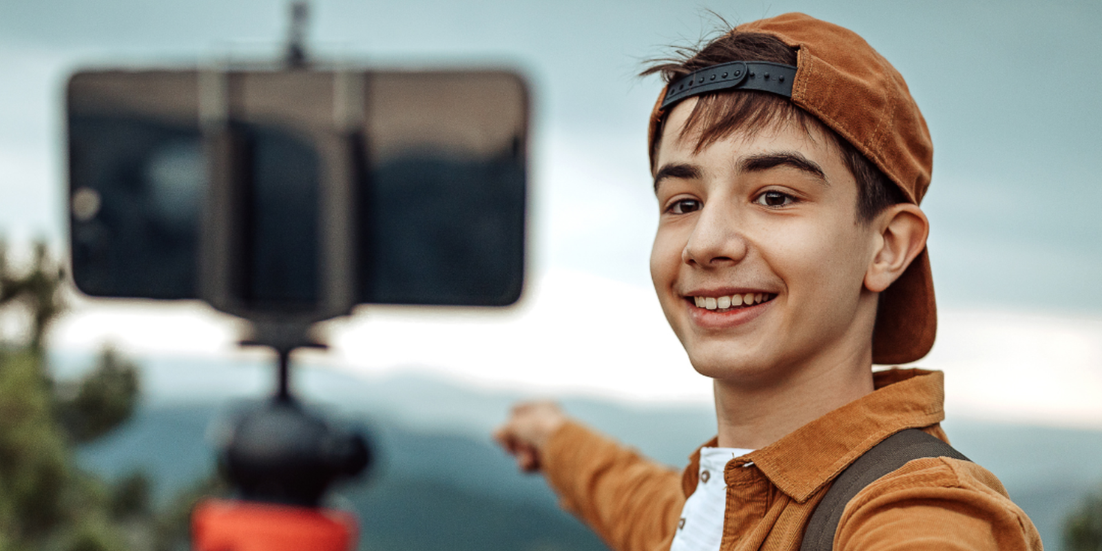 A young boy wearing a cap vlogs with a smartphone on a selfie stick, set against a mountainous backdrop.