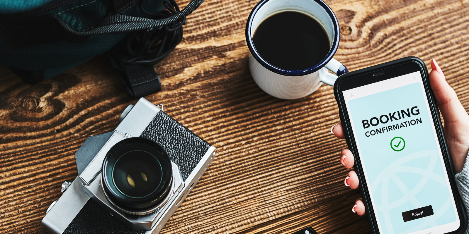 A person's hand holding a smartphone displaying a "Booking Confirmation" screen, with a camera and a cup of coffee on a wooden table, indicating travel preparation.